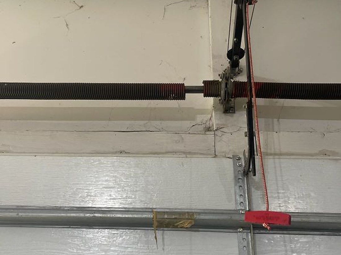 Torsion springs and shaft mounted across the header above a closed residential garage door, cable drums and red release cord in view