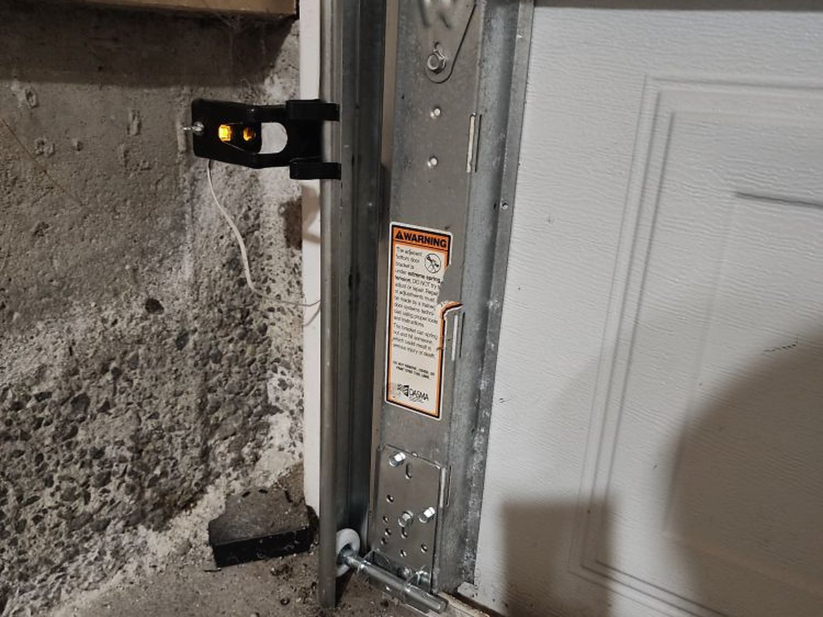 Base of a vertical garage door track with the bottom roller bracket and lift cable, and the wall-mounted safety sensor beside the opening